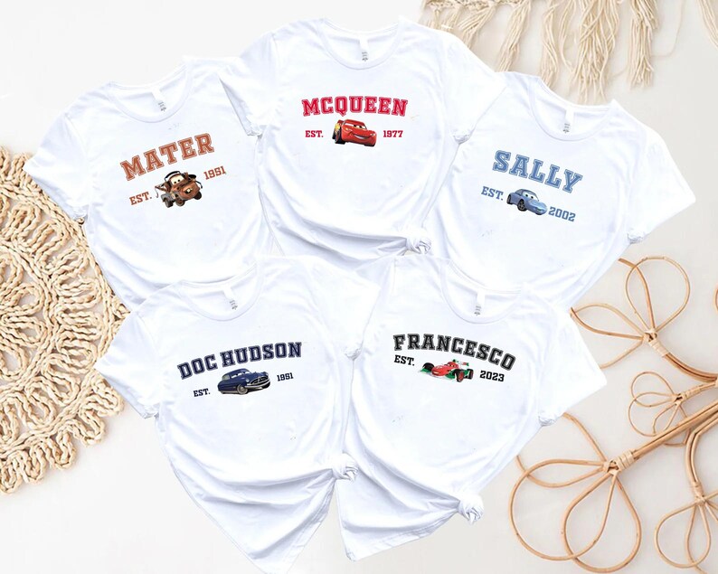 May include: White t-shirts with character names and car graphics from the film Cars. Each shirt has a character's name, a car image, and the year of their introduction. Characters include Mater, McQueen, Sally, Doc Hudson, and Francesco.