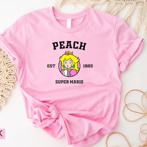 May include: Pink t-shirt with a peach character graphic and the text "Peach Est. 1985 Super Mario".