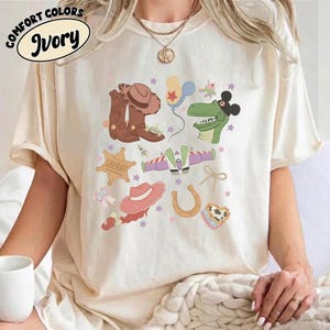 May include: Ivory-colored t-shirt with a colorful graphic featuring cowboy boots, a sheriff badge, a horseshoe, and other playful illustrations. The shirt has the words "Comfort Colors" and "Ivory" printed on it.
