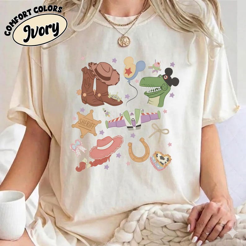 May include: Ivory-colored t-shirt with a colorful graphic featuring cowboy boots, a sheriff badge, a horseshoe, and other playful illustrations. The shirt has the words "Comfort Colors" and "Ivory" printed on it.