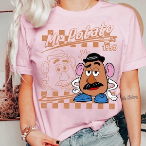 Może przedstawiać: Jasnor&oacute;żowy t-shirt z kr&oacute;tkim rękawem z nadrukiem graficznym przedstawiającym Mr. Potato Head. Wz&oacute;r zawiera napis Mr. Potato 1995, brązowe tło w kratkę oraz kolorową ilustrację postaci w czarnym kapeluszu i niebieskich butach.