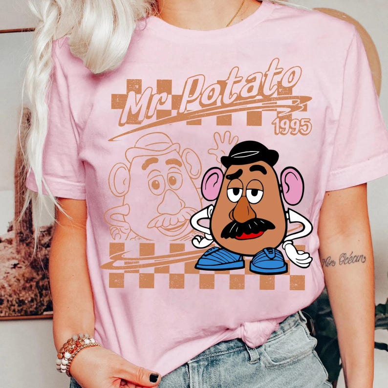 Może przedstawiać: Jasnor&oacute;żowy t-shirt z kr&oacute;tkim rękawem z nadrukiem graficznym przedstawiającym Mr. Potato Head. Wz&oacute;r zawiera napis Mr. Potato 1995, brązowe tło w kratkę oraz kolorową ilustrację postaci w czarnym kapeluszu i niebieskich butach.