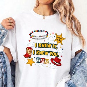 May include: A white t-shirt featuring a colorful graphic print with the text "I KNEW IT, I KNEW YOU," a beaded bracelet with the text "YOU'VE GOT A FRIEND IN ME," a yellow sheriff star with "INFINITY AND BEYOND," cowboy boots, blocks, and a hat.