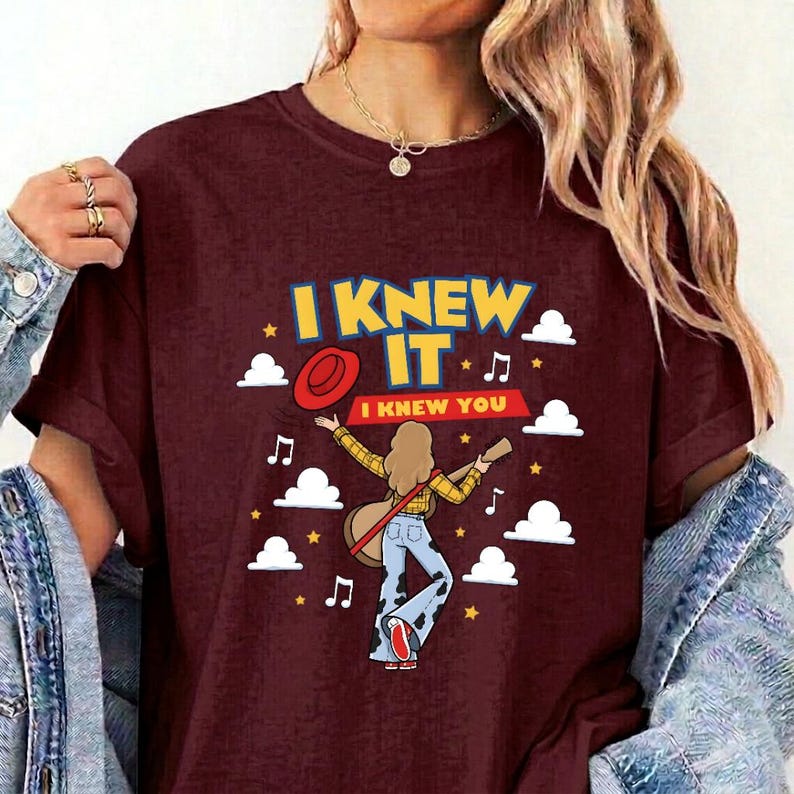 May include: A maroon short-sleeve t-shirt featuring a graphic of a person with long hair playing a guitar, wearing a yellow plaid shirt and cow-print pants. The text reads I KNEW IT and I KNEW YOU, surrounded by white clouds, stars, and musical notes.