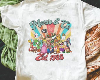 Peut inclure: Un t-shirt blanc à manches courtes avec une impression graphique colorée représentant divers personnages de jeux vidéo. Le texte indique Mario & Co. Est. 1985, sur un fond de rayures en éventail dans des tons rouge, orange, jaune et bleu.