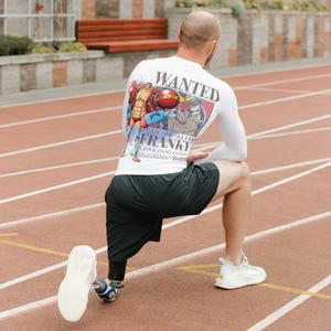 Puede incluir: Camiseta blanca de manga larga con un diseño de póster de "SE BUSCA" en la espalda, con personajes de dibujos animados y el nombre "FRANKY". La persona lleva pantalones cortos negros y zapatillas blancas, arrodillada en una pista roja.
