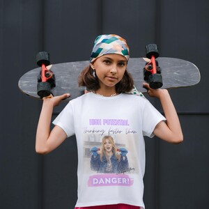 May include: A person holding a skateboard, wearing a white t-shirt with the text "HIGH POTENTIAL thinking faster than DANGER!". The person is also wearing a patterned headscarf.