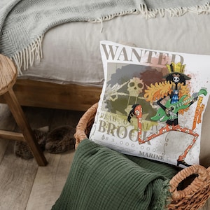 May include: White cushion featuring a cartoon skeleton playing a green guitar. The cushion has the text "WANTED" and "DEAD OR ALIVE BROOK". The cushion is in a wicker basket with a green blanket.