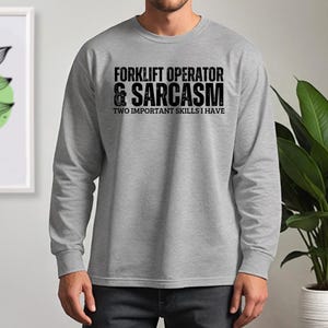 May include: A gray long-sleeve shirt with the text "FORKLIFT OPERATOR & SARCASM TWO IMPORTANT SKILLS I HAVE" printed in black. The shirt is a crew neck style.