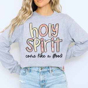 May include: A light gray long-sleeve shirt with the words "holy spirit" in a floral and checkered design. The phrase "come like a flood" is printed below the words. The shirt is made of a soft material.
