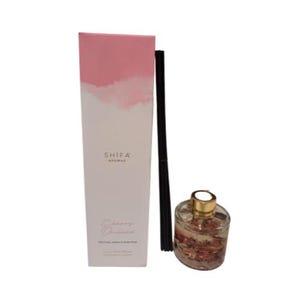 Puede incluir: Una caja rosa y blanca con el texto "SHIFA AROMAS" y "Cherry Orchard" en ella. La caja está al lado de una botella de vidrio transparente con una tapa dorada y palitos negros. La botella está llena de un líquido transparente y tiene pequeñas flores rosas y blancas en su interior.