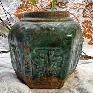 May include: A vintage, green ceramic jar with a textured surface and embossed floral designs. The jar has a wide, rounded body and a narrow, slightly flared rim. The color is a mix of green and brown, with a weathered appearance. The jar is a decorative item.