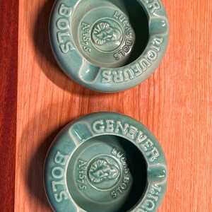 May include: Two round, light green ceramic ashtrays featuring embossed text that reads GENEVER, BOLS, and LIQUEURS. The center of each ashtray displays a circular emblem with the text LUCAS BOLS and Aº 1575, along with a small relief portrait.