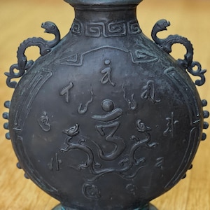 May include: Dark bronze-colored vase with a round body and a narrow neck. The vase features embossed Asian-inspired symbols and dragon-shaped handles. The base is circular, and the overall design suggests an antique or decorative item.