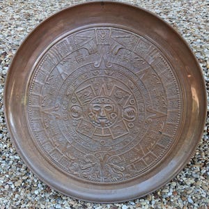May include: A circular bronze-toned metal tray featuring a detailed embossed relief of the Aztec sun stone calendar. The central design displays a human face surrounded by concentric rings of intricate geometric patterns and symbols.
