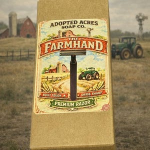 May include: A brown cardboard box featuring a vintage-style label for "The Farmhand Premium Razor" by Adopted Acres Soap Co. The label depicts a rural scene with a tractor, barn, and the text "Built Tough" and "Shave Sharp".