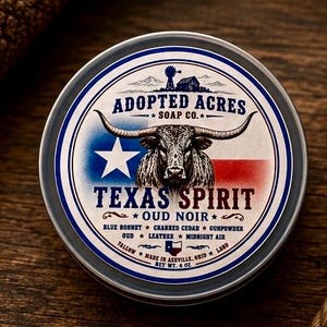 May include: A round metal tin of Adopted Acres Soap Co. Texas Spirit Oud Noir soap. The label features a longhorn steer, a Texas flag design, and text listing scent notes including blue bonnet, charred cedar, gunpowder, oud, leather, and midnight air. Net weight 4 oz.