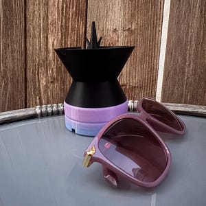 May include: A black and purple object with a conical top and a cylindrical base, next to a pair of pink sunglasses with gold accents. The sunglasses are open, displaying the lenses. The background is a wooden wall.