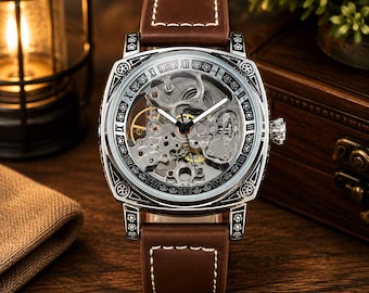 Puede incluir: Un reloj de pulsera mecánico de caja cuadrada con correa de cuero marrón y costuras blancas. La esfera muestra un movimiento esqueleto con engranajes visibles, manecillas plateadas y un bisel negro decorado con números romanos y motivos florales.