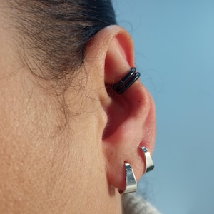 Peut inclure: Gros plan sur une oreille ornée de deux boucles d'oreilles créoles argentées et d'un ear cuff noir. Le ear cuff présente un design à double bande. Les boucles d'oreilles sont de style simple et minimaliste. La peau est claire, et le fond est bleu clair.