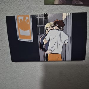 May include: A print of an illustration depicting two figures embracing. One figure is wearing a white shirt and orange pants, while the other is in a black shirt. A framed image with an orange design hangs on the wall.