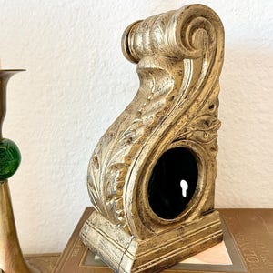 May include: A gold-toned decorative wall bracket with intricate carvings and a keyhole opening. The bracket is sitting on a stack of books.