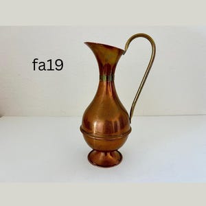 May include: A vintage copper pitcher with a brass handle. The pitcher has a bulbous body and a flared spout. The handle curves gracefully from the top to the base. The pitcher is likely used for decorative purposes.