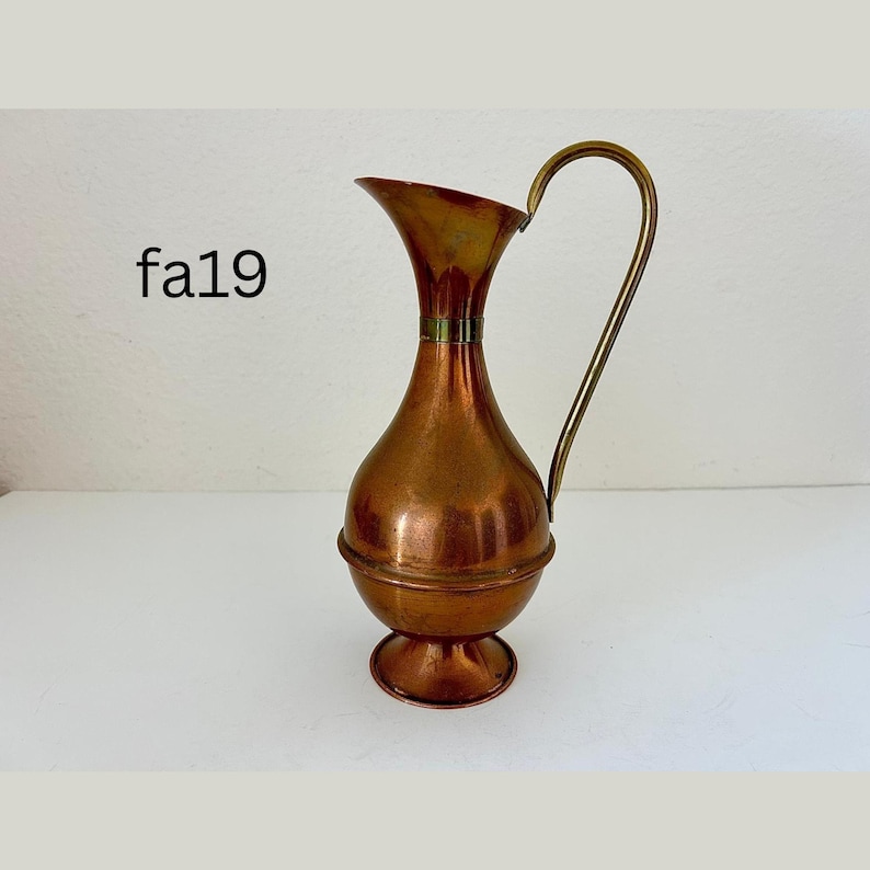May include: A vintage copper pitcher with a brass handle. The pitcher has a bulbous body and a flared spout. The handle curves gracefully from the top to the base. The pitcher is likely used for decorative purposes.