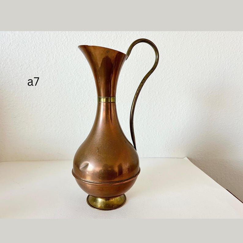 May include: A copper-colored pitcher with a brass handle and base. The pitcher has a bulbous body and a flared top. The handle curves gracefully from the top to the side. The text "a7" is visible on the left side.