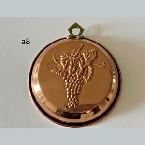 May include: A round, copper-colored wall hanging with a grape vine design. The decorative piece has a raised relief of grapes and leaves. It features a small brass loop at the top for hanging. The text "a8" is in the upper left corner.
