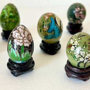 May include: A collection of decorative, hand-painted egg-shaped ornaments. Each egg features unique floral and bird designs in various colors, set on small, dark wooden stands. The eggs showcase intricate details and vibrant hues, creating a charming display.