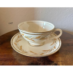 May include: Cream-colored teacup and saucer set with gold trim and wheat stalk design. The cup has a handle and sits on a matching saucer. The set is vintage and likely made of porcelain.