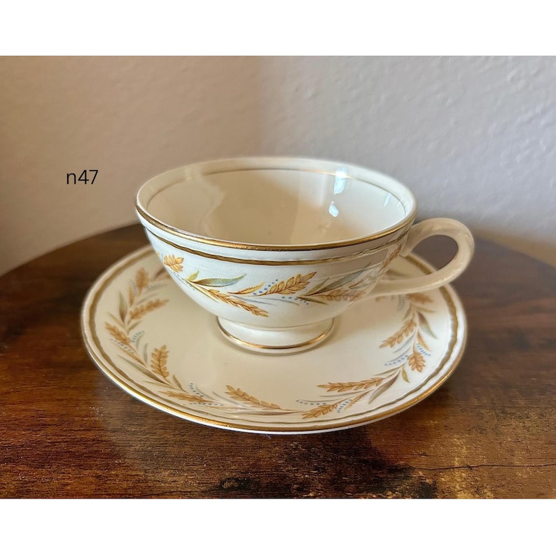 May include: Cream-colored teacup and saucer set with gold trim and wheat stalk design. The cup has a handle and sits on a matching saucer. The set is vintage and likely made of porcelain.