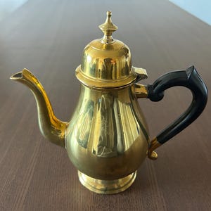 May include: A vintage brass teapot with a shiny gold finish. It features a black handle and a decorative finial on the lid. The teapot is designed with a curved spout and sits on a round base, perfect for serving tea or coffee.