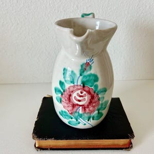 May include: A white ceramic pitcher with a floral design. The pitcher features a red and pink rose with green leaves and blue accents. The handle is a small, curved shape. The pitcher is sitting on a black book.