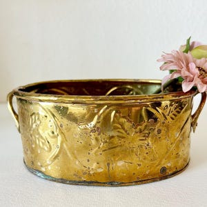 May include: A gold-toned brass planter with ornate floral designs and two handles. The planter is empty and has a worn, antique look.