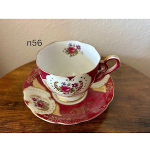 May include: A vintage teacup and saucer set. The teacup and saucer are white with a red border and gold trim. Floral designs decorate the cup and saucer. The teacup has a curved handle. The text "n56" is visible.
