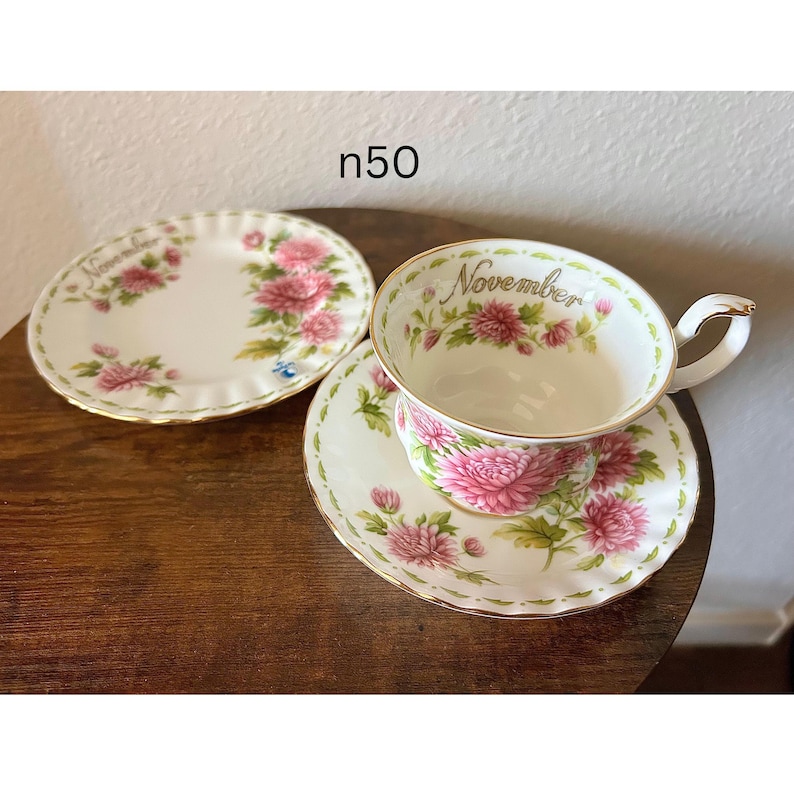 May include: A vintage teacup, saucer, and side plate set. The white porcelain pieces feature pink floral designs and gold trim. The teacup is labeled "November". The set is displayed on a wooden surface.