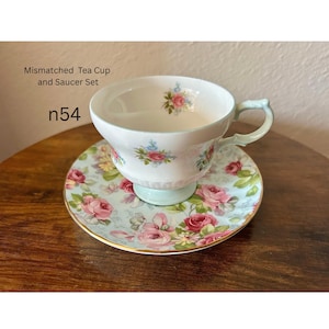 May include: A mismatched tea cup and saucer set. The cup and saucer are decorated with pink roses and blue accents on a light blue background. The cup has a light green handle and base. The text "Mismatched Tea Cup and Saucer Set" and "n54" are visible.