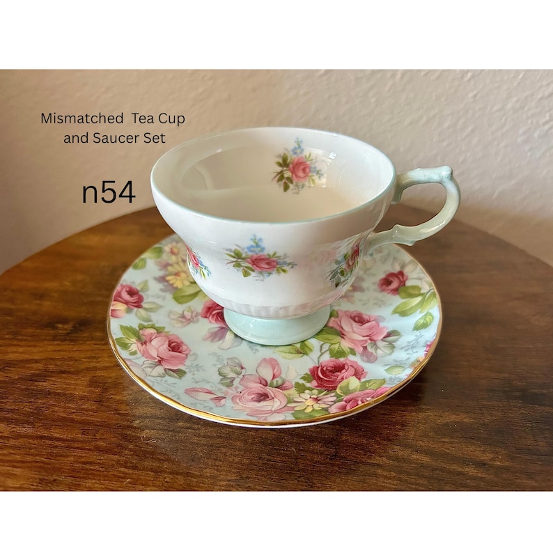 May include: A mismatched tea cup and saucer set. The cup and saucer are decorated with pink roses and blue accents on a light blue background. The cup has a light green handle and base. The text "Mismatched Tea Cup and Saucer Set" and "n54" are visible.