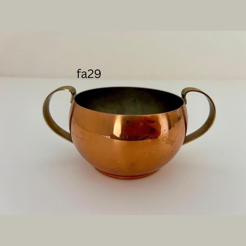 May include: A copper bowl with two brass handles. The bowl has a rounded shape and a polished finish, reflecting light. The text "fa29" is printed on the top left of the image. This item is likely vintage or antique.