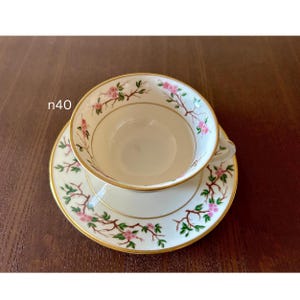 May include: A white teacup and saucer set with a floral design. The set features pink blossoms and green leaves on delicate branches, with gold trim. The teacup sits on the matching saucer, both on a dark brown surface. The text "n40" is visible.