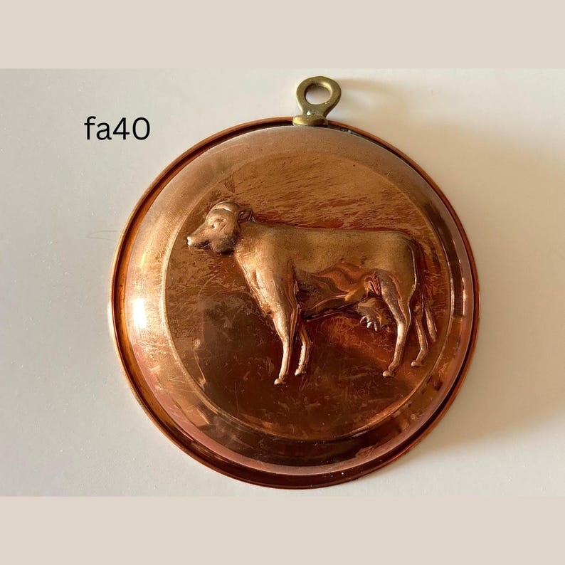May include: A copper-colored, round mold with a cow relief. The mold has a brass loop for hanging and the text "fa40" on the left side. The cow is depicted in profile, with detailed features.