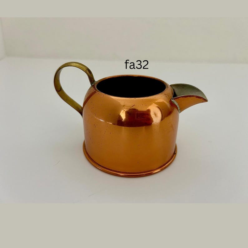May include: A small, copper-colored creamer with a brass handle and a spout. The creamer has a round body and a dark interior. The text "fa32" is visible on the upper right side of the image.