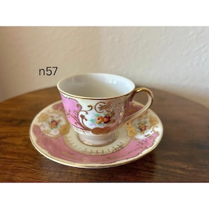 May include: A vintage teacup and saucer set. The cup and saucer are decorated with pink, gold, and floral designs. The cup has a gold handle and rim. The saucer has a matching pink and gold border. The text "n57" is visible.