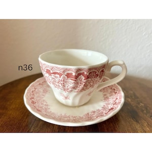 May include: A white teacup and saucer set with a red floral pattern. The teacup has a curved handle and sits on a matching saucer. The set is on a wooden surface. The text "n36" is visible.