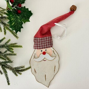 May include: A wooden Santa Claus Christmas ornament with a red hat and a white beard. The hat has a plaid pattern at the base. A small bell is attached to the end of the hat. The ornament is hanging by a white string.