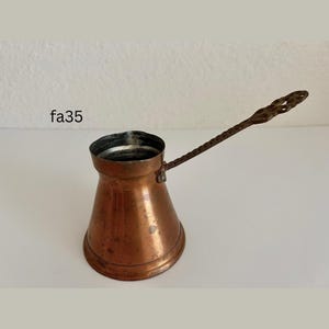 May include: A copper-colored Turkish coffee pot with a long, twisted handle. The pot has a wide base that tapers to a narrow neck and a flared rim. The handle is attached to the side of the pot. The text "fa35" is visible in the image.