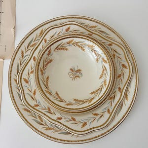 May include: A set of three stacked ceramic dishes featuring a cream-colored base with a scalloped edge. Each piece is decorated with a repeating pattern of golden-brown wheat stalks and subtle grey leaves, centered with a wheat motif.