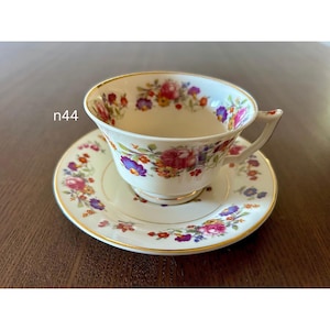 May include: A vintage teacup and saucer set. The white porcelain cup and saucer are adorned with a floral pattern in shades of red, purple, yellow, and blue. The rims are trimmed with gold. The text "n44" is visible on the side.